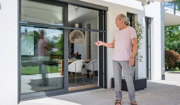 Älterer Mann mit rosa T-shirt und Jeans steht aussen vor einer Terassentür und bedient mit einer Fernbedienung ein Insektenschutz Elektrorollo