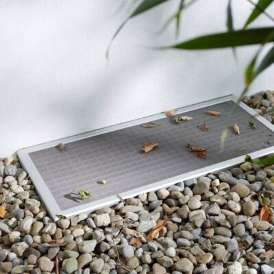 eine Aluminium Lichtschachtabdeckung mit Rahmen und Edelstahlgewebe liegt auf einem Gitterrost an der Hauswand und ist von Kies umgeben.