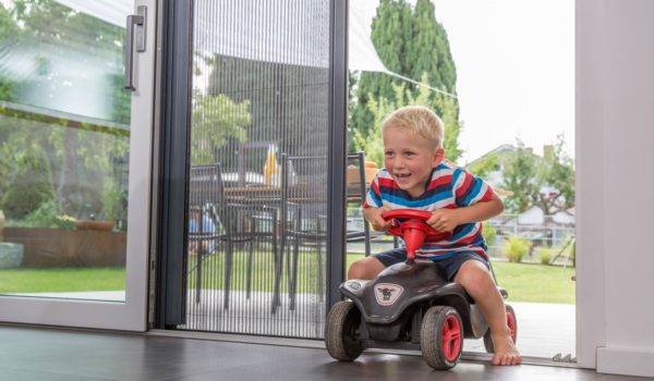 blondes Kind ca. 3 Jahre alt mit bunt gestreiften Shirt sitzt auf einem Spielzeugauto und fährt durch eine offene Tür mit Fliegengitterplissee