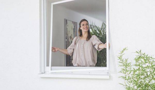 Frau mit dunklen langen Haaren und beiger Bluse steht innen am Fenster und hängt einen Fliegengitter Spannrahmen am Fenster ein