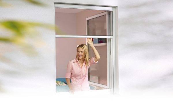 Junge Frau mit langen blonden Haaren und rosa Bluse steht innen am Fenster und öffnet ein Insektengitterrollo