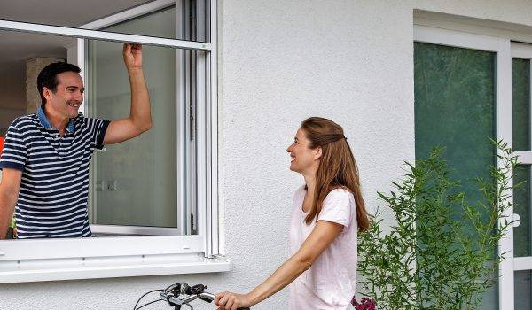 Mann steht innen am geöffneten Fenster und schiebt ein Insektenschutzgitter Rollo nach oben. Dabei unterhält er sich mit einer Frau welche aussen vor dem Fenster steht und ein Fahrrad in der Hand hält