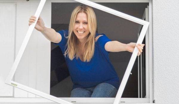 Sara Bachmann, eine Frau mit blonden Haaren und blauem Shirt hält einen Fliegengitter Spannrahmen durch ein offenes Fenster uns lacht dabei