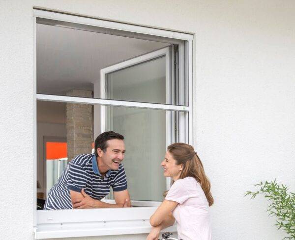 Ein Mann und eine Frau unterhalten sich durch ein offenes Fenster. Das Insektenschutzrollo ist halb aufgerollt.