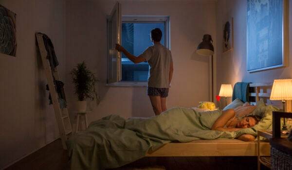 Frau liegt im Bett und Mann öffnet das Fenster mit Fliegengitter